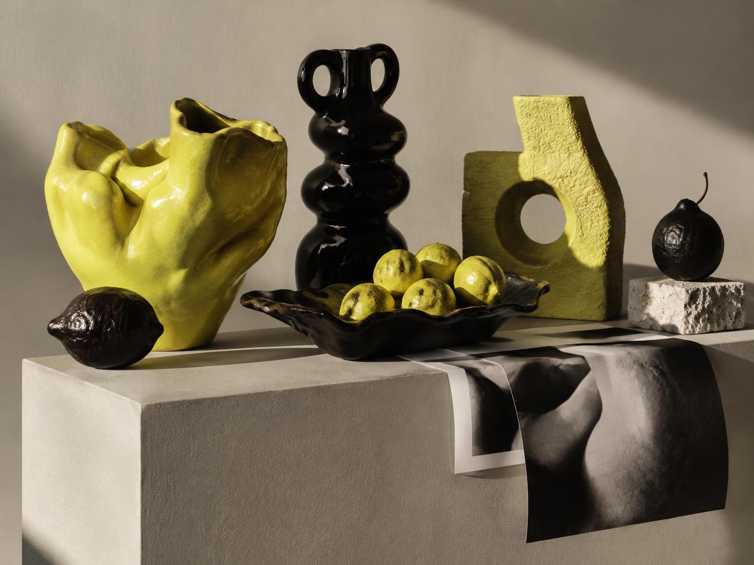 Project Nova — a still-life arrangement of chartreuse and glossy black ceramic vessels, a bowl of yellow citrus, and a draped photographic print on a pale stone plinth